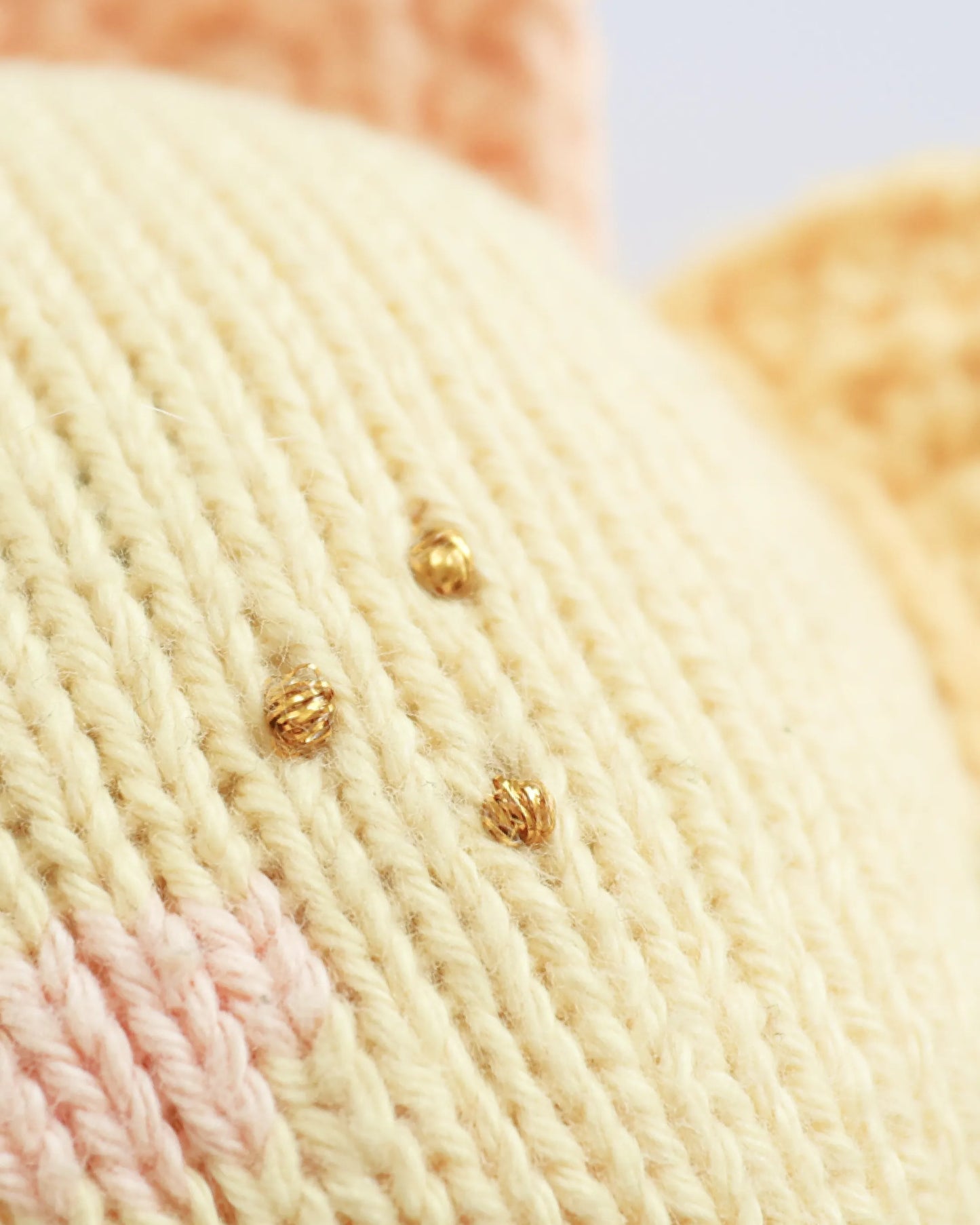Close-up of a cream-colored knitted textile with small metallic gold bead accents and a soft pink section, shown in high detail.