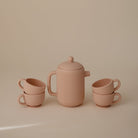 Silicone Tea Play Set by Mushie & Co in blush pink, featuring a lidded teapot and three matching tea cups arranged on a neutral background.