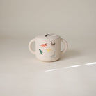 White children's cup with dinosaur illustrations on a beige background