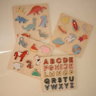 A flatlay arrangement of four wooden puzzles on a white surface, including a dinosaur puzzle, a space-themed puzzle, a farm animal puzzle, and an alphabet puzzle featuring red wooden letters.