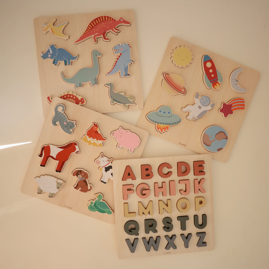A flatlay arrangement of four wooden puzzles on a white surface, including a dinosaur puzzle, a space-themed puzzle, a farm animal puzzle, and an alphabet puzzle featuring red wooden letters.