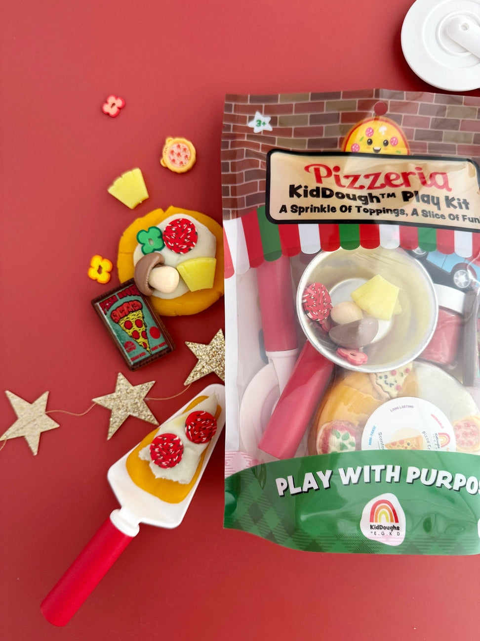Pizzeria KidDough play kit in its packaging, displayed on a red background with toy pizza slices, a spatula, and star decorations.