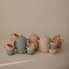A collection of silicone tea play sets by Mushie & Co featuring teapots, lids, and matching cups in sage green and blush pink, arranged on a neutral surface.