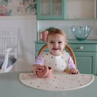 Child sitting at a table with a polka dot placemat and bib, holding a pink bowl.