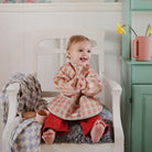 Child in a colorful outfit sitting on a white chair in a room with a green cabinet and pink mug.