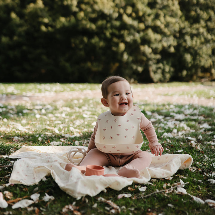 Baby sitting on a blanket in a park with greenery in the background
