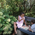 A young girl sitting outdoors on a bench holding a piece from a wooden puzzle, with another toddler sitting nearby looking at a wooden puzzle placed on the bench surface.