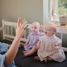 Two babies sitting on a bed with a person's hand reaching out, likely playing with them.