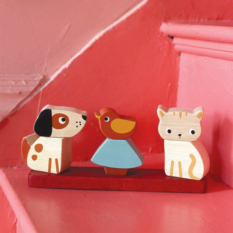 Wooden Pet Stacker toy by Mentari featuring a rectangular red base holding three removable pieces: a spotted white and brown puppy on the left, a blue and yellow bird on a tree in the center, and a white kitten with orange markings on the right, all resting on a pink surface against a soft, matching pink background.