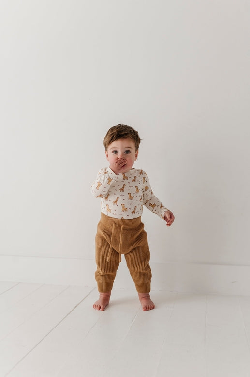 A toddler standing barefoot in a bright white room, wearing a cream long-sleeve top with a small brown animal print and brown corduroy pants.