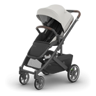 A premium stroller shown at a three-quarter angle on a clean white background. The stroller features a light beige/cream canopy, dark gray frame, brown leatherette handle grips, and a black seat liner.