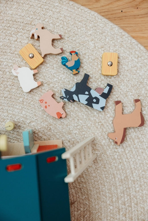 An overhead, bird's-eye view of the Wooden Big Red Barn Playset by Mentari components laid out on a textured surface, including seven wooden animal figures—a cow, horse, pig, sheep, goat, chicken, and rooster—alongside a blue ladder, a small stool, and a piece of fencing.