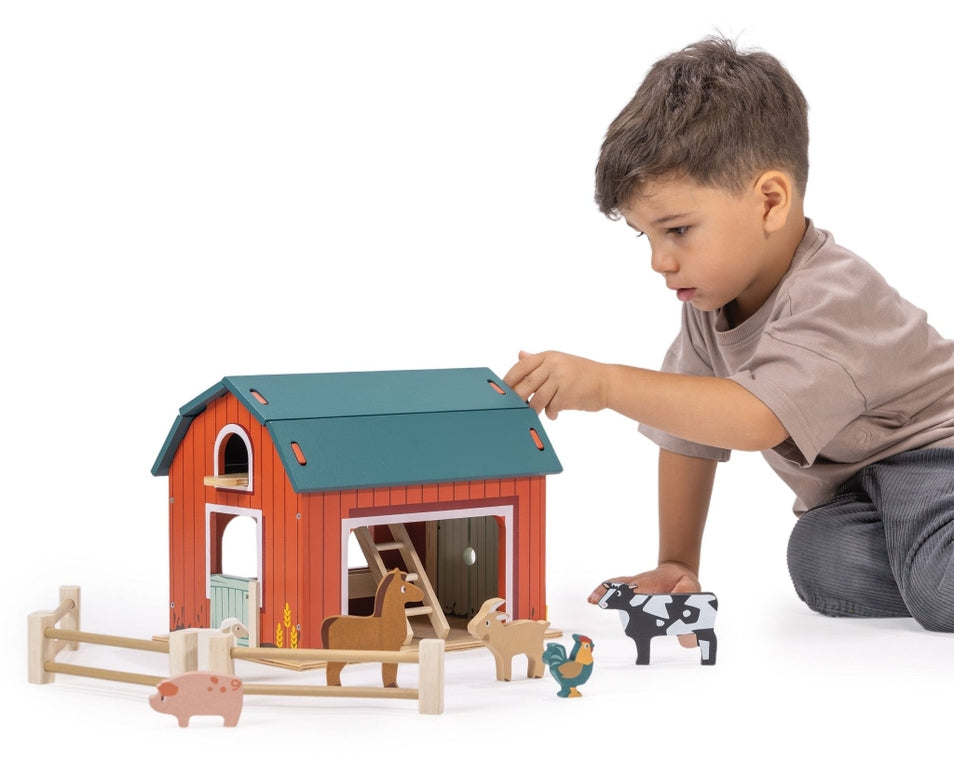 A young boy with short brown hair, wearing a gray t-shirt and jeans, plays with the Wooden Big Red Barn Playset by Mentari, reaching for the roof of the red barn while wooden farm animal figures stand in the fenced enclosure below.