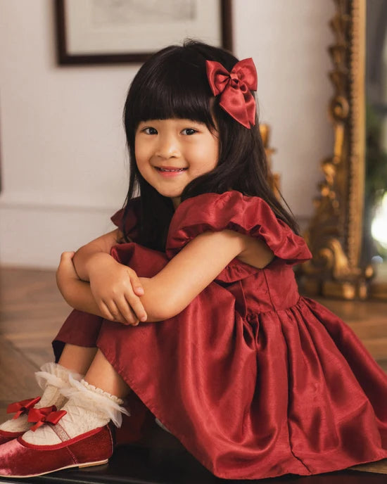A young girl with dark hair wearing a shiny red satin dress, a red bow in her hair, and red shoes with white tulle trim, sitting in an indoor setting.