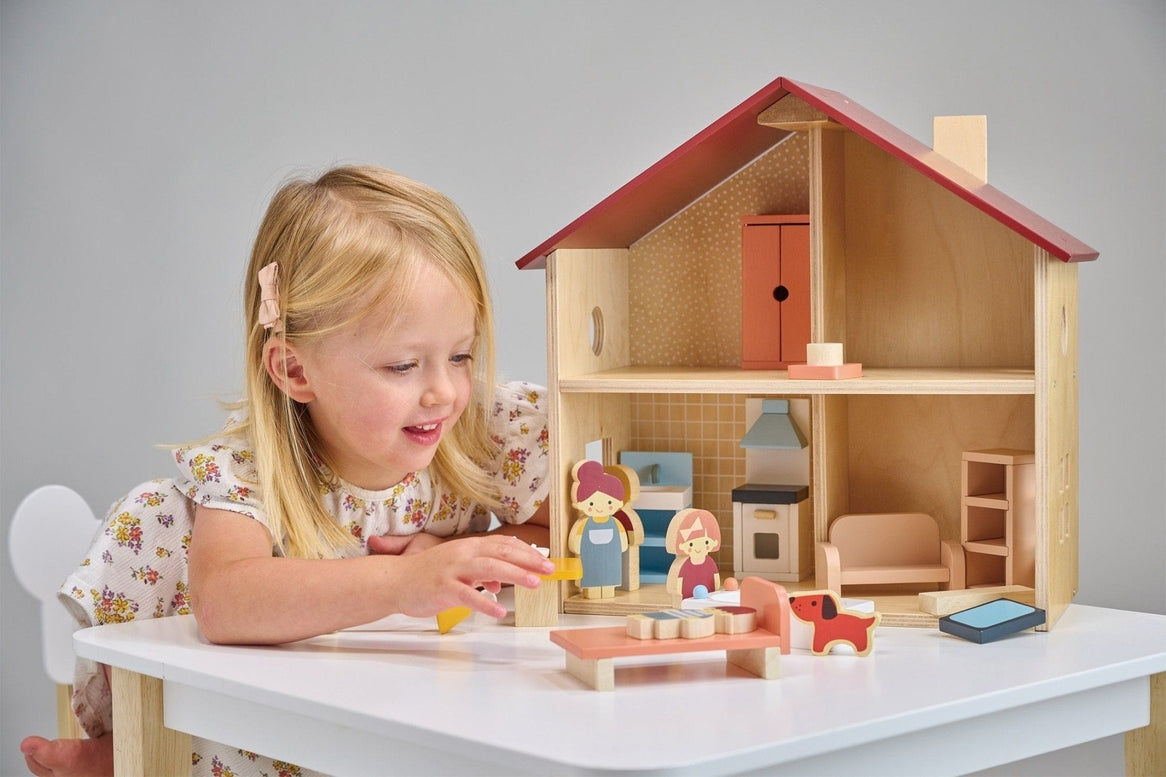 A young child playing with an open-faced wooden dollhouse by Mentari, arranged on a white table with miniature wooden furniture and small figurines.
