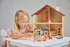 A young child playing with an open-faced wooden dollhouse by Mentari, arranged on a white table with miniature wooden furniture and small figurines.