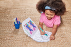 A young girl with an afro hairstyle, wearing a pink shirt and blue headband, is sitting on a beige woven rug and coloring a book with a green marker. A small blue cup filled with colored pencils is next to her.