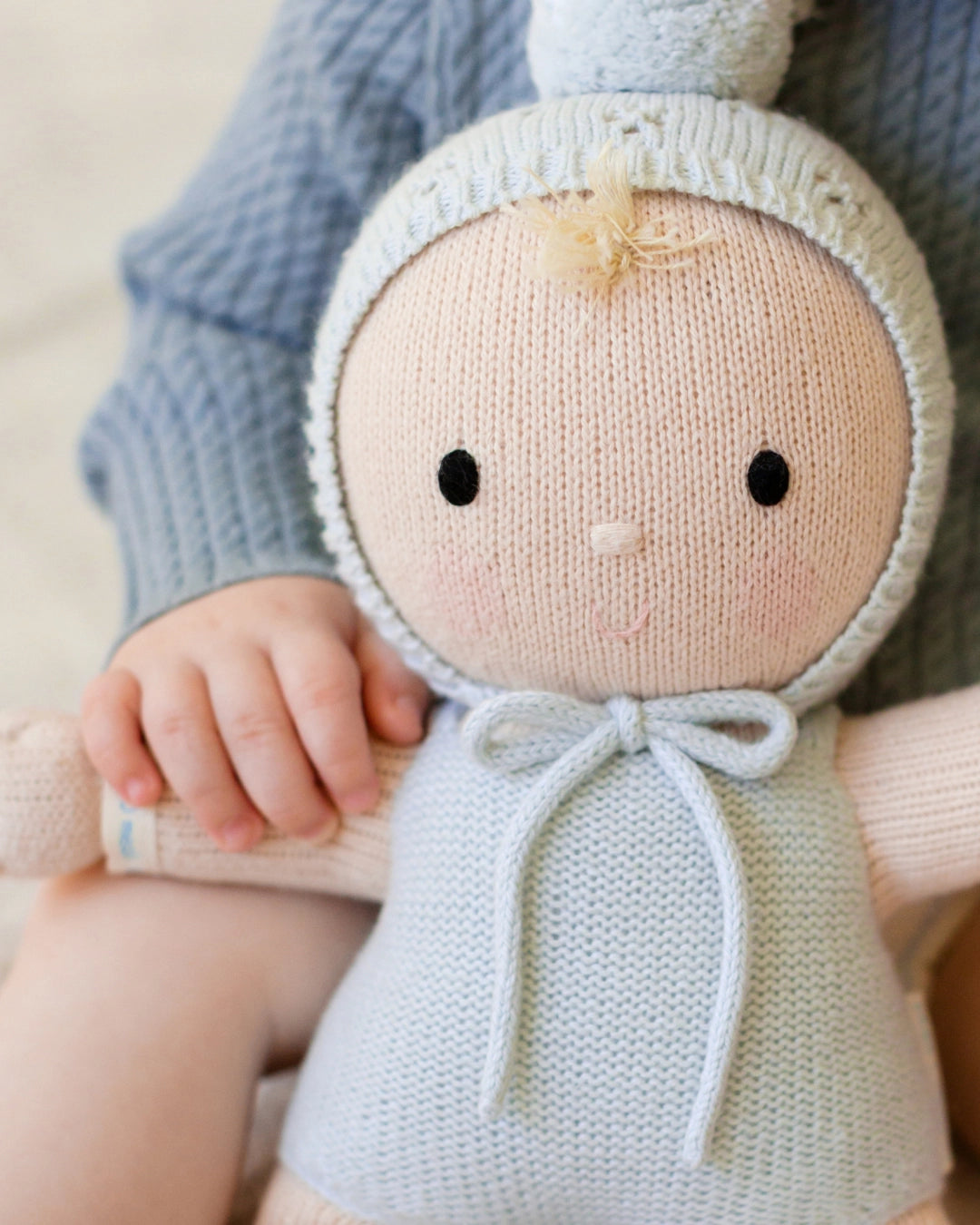 A close-up of a knitted doll being held by a person wearing a blue cable-knit sweater. The doll has a pale peach face and hands, a light blue knit bonnet and bodysuit, and simple black embroidered eyes.