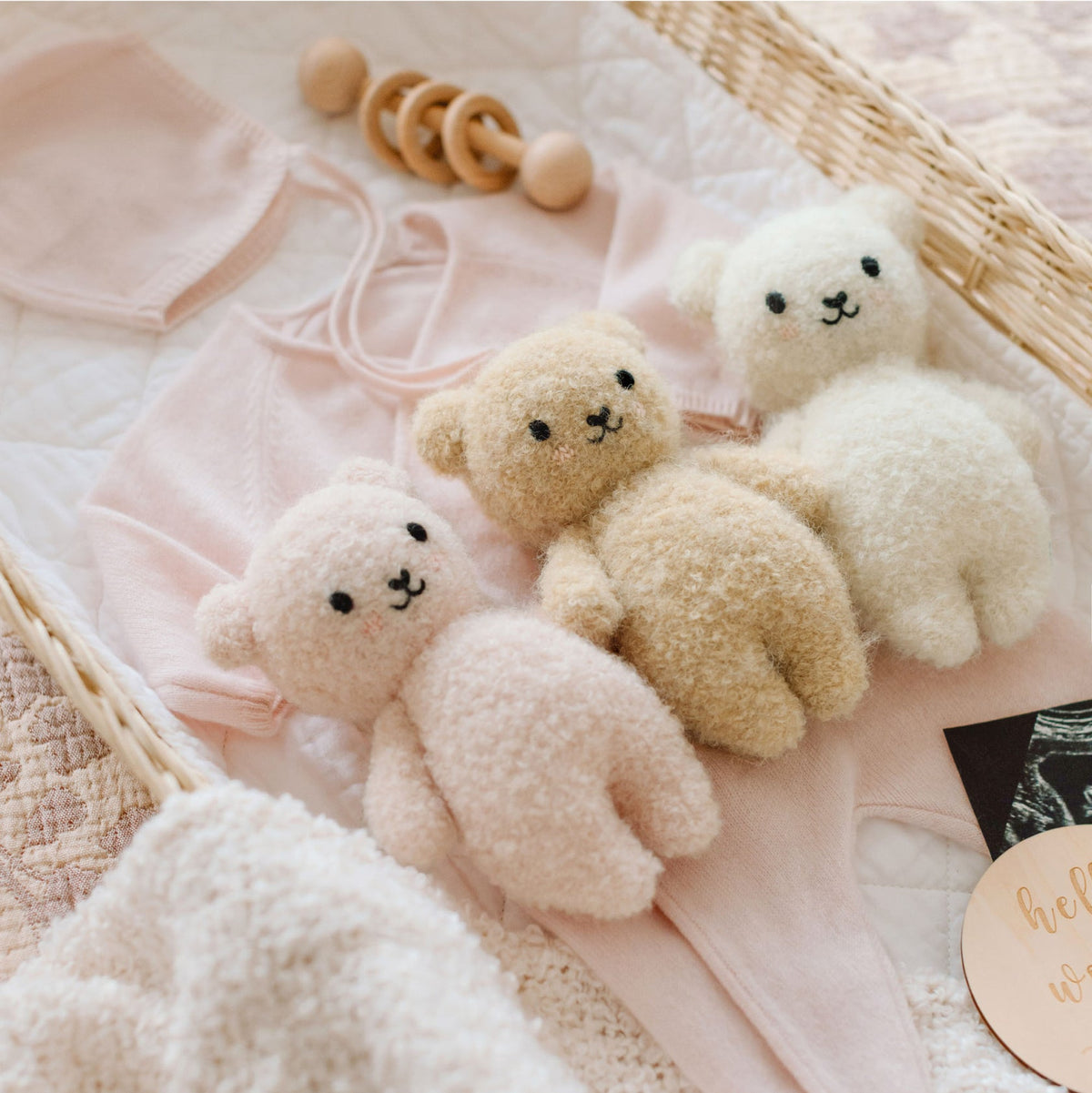 Three small teddy bears on a pink blanket with wooden toys and a heart-shaped object.