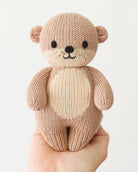A small knitted teddy bear held in a person's hand against a plain light background. The bear is made from soft knit fabric in warm beige and tan tones with black embroidered eyes, nose, and mouth.