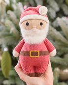 A knitted Santa Claus held in a person's hand doll wearing a pink and white hat with a pom-pom, a white beard, and a pink outfit with a brown belt and boots, isolated on a green background.