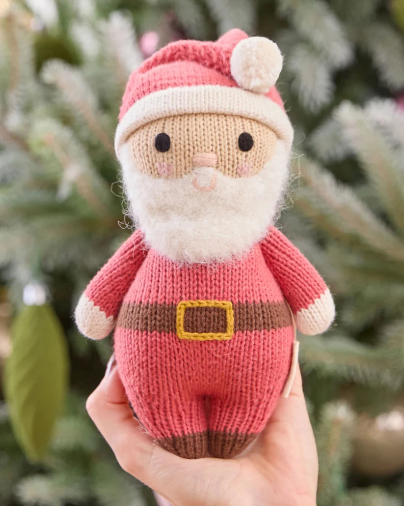 A knitted Santa Claus held in a person's hand doll wearing a pink and white hat with a pom-pom, a white beard, and a pink outfit with a brown belt and boots, isolated on a green background.