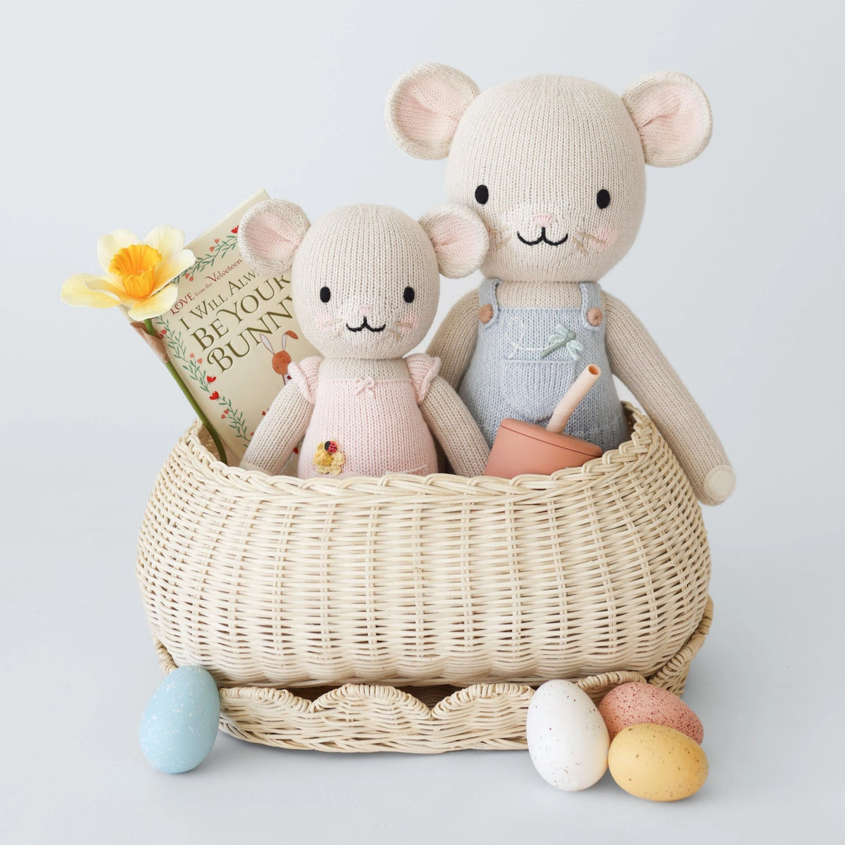 Two knitted mouse toys in a wicker basket with a book and Easter eggs on a light gray background