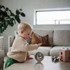 A toddler wearing a beige knit sweater sitting on a living room floor next to a pink toy block, playing with a Mushie Suction Spinner Ball.