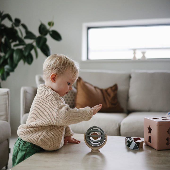A toddler wearing a beige knit sweater sitting on a living room floor next to a pink toy block, playing with a Mushie Suction Spinner Ball.