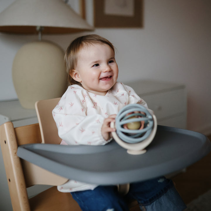 A smiling baby sitting in a wooden high chair, looking at and touching a blue and beige Mushie Suction Spinner Ball attached to the tray.