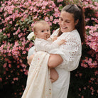 smiling woman holding a baby wrapped in a white smiley-face muslin swaddle blanket outdoors in front of pink flowers.