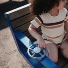 Child sitting on a blue seesaw with a steering wheel, wearing a striped shirt and shorts.