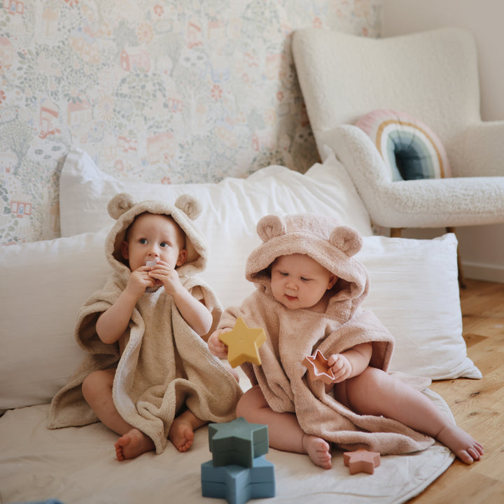 Two children in bear-themed hooded towels sitting on a bed with toys around.