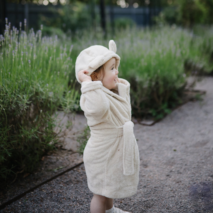 Child in a cream-colored robe and hat standing in a garden with lavender plants.