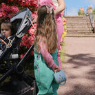Two children, one in a stroller and the other holding a cup, walking along a path with pink flowers and steps in the background.