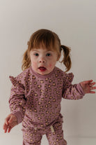 A toddler girl with brown hair in pigtails wearing a mauve pajama set with a leopard print pattern, standing against a plain light gray background.