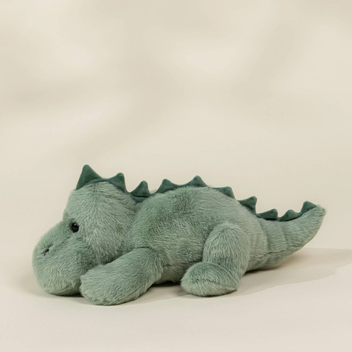 A plush dinosaur toy in a dusty sage green color, lying down on a plain light beige background.