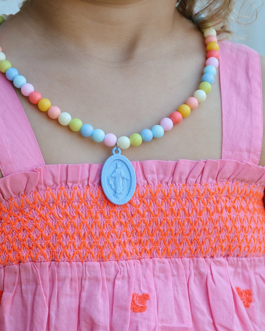 Child wearing a colorful beaded necklace with a blue pendant on a pink dress.