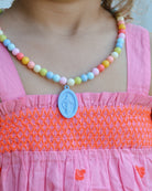 Child wearing a colorful beaded necklace with a blue pendant on a pink dress.