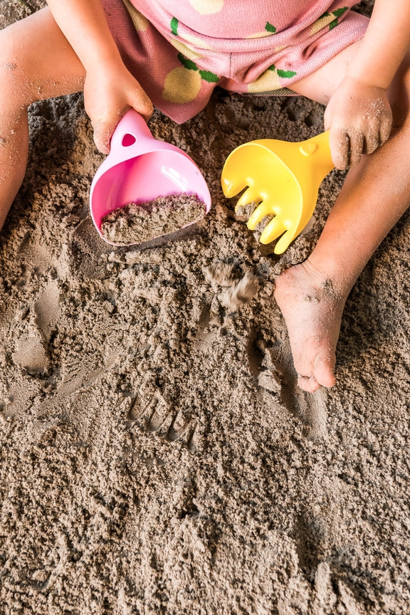 A child sitting in the sand playing with a pink digger toy and a yellow rake toy.