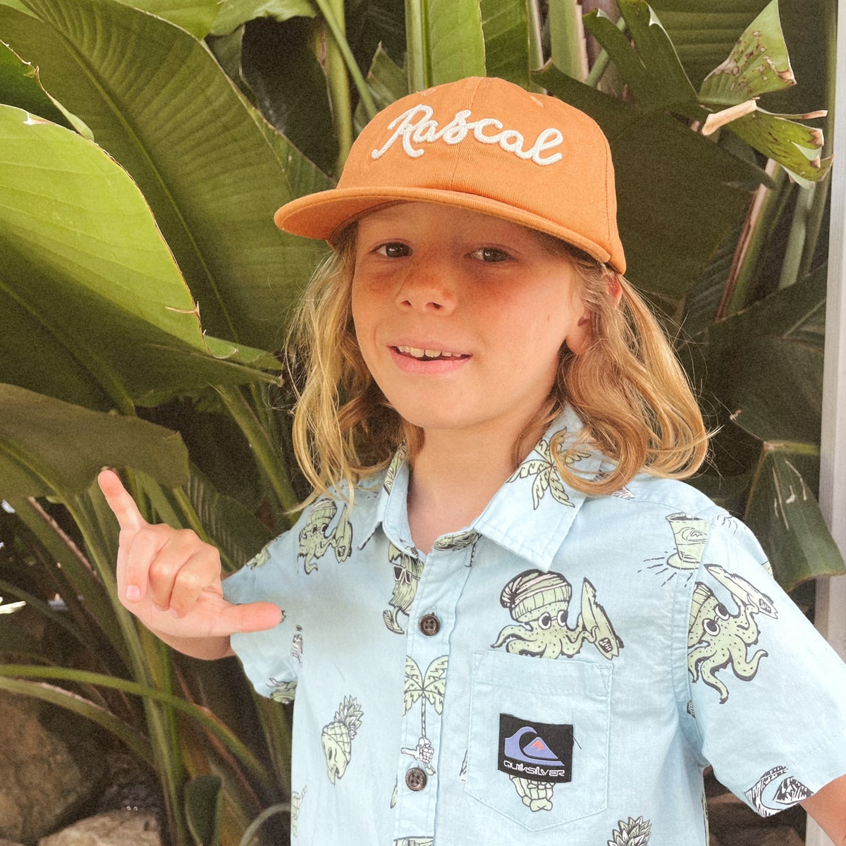 A young boy wearing a light blue Quiksilver short-sleeve button-up shirt with a dinosaur print and a tan baseball cap with 'Rascal' embroidered on it, posing in front of large green tropical leaves.