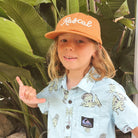 A young boy wearing a light blue Quiksilver short-sleeve button-up shirt with a dinosaur print and a tan baseball cap with 'Rascal' embroidered on it, posing in front of large green tropical leaves.