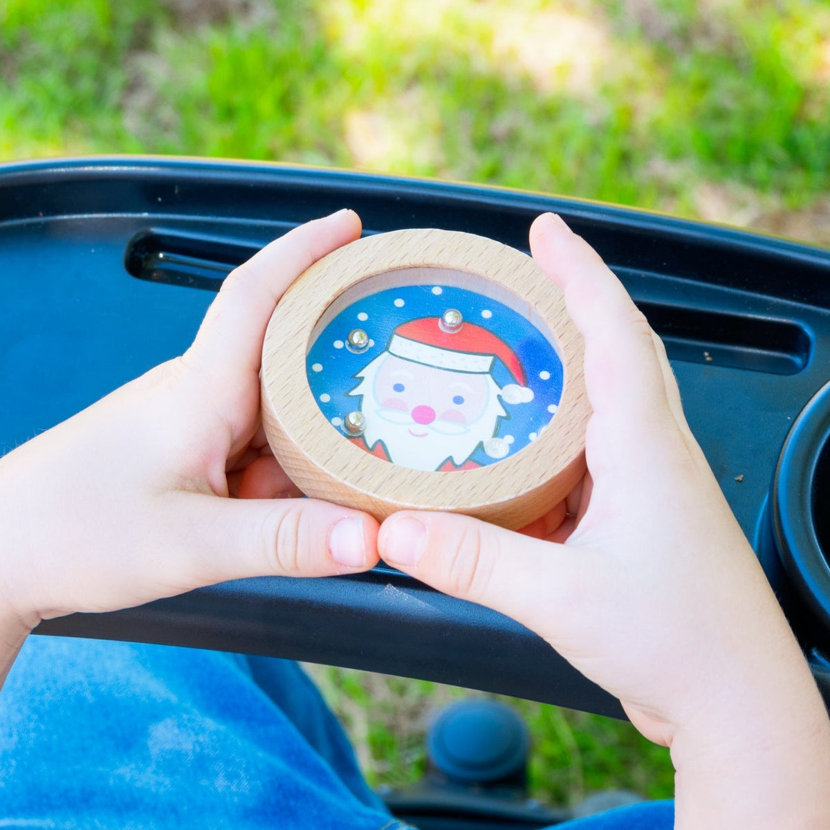 A child's hands holding a round wooden toy container with a Santa Claus illustration, positioned over a black stroller tray in an outdoor setting with green grass in the background.