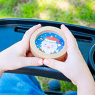 A child's hands holding a round wooden toy container with a Santa Claus illustration, positioned over a black stroller tray in an outdoor setting with green grass in the background.