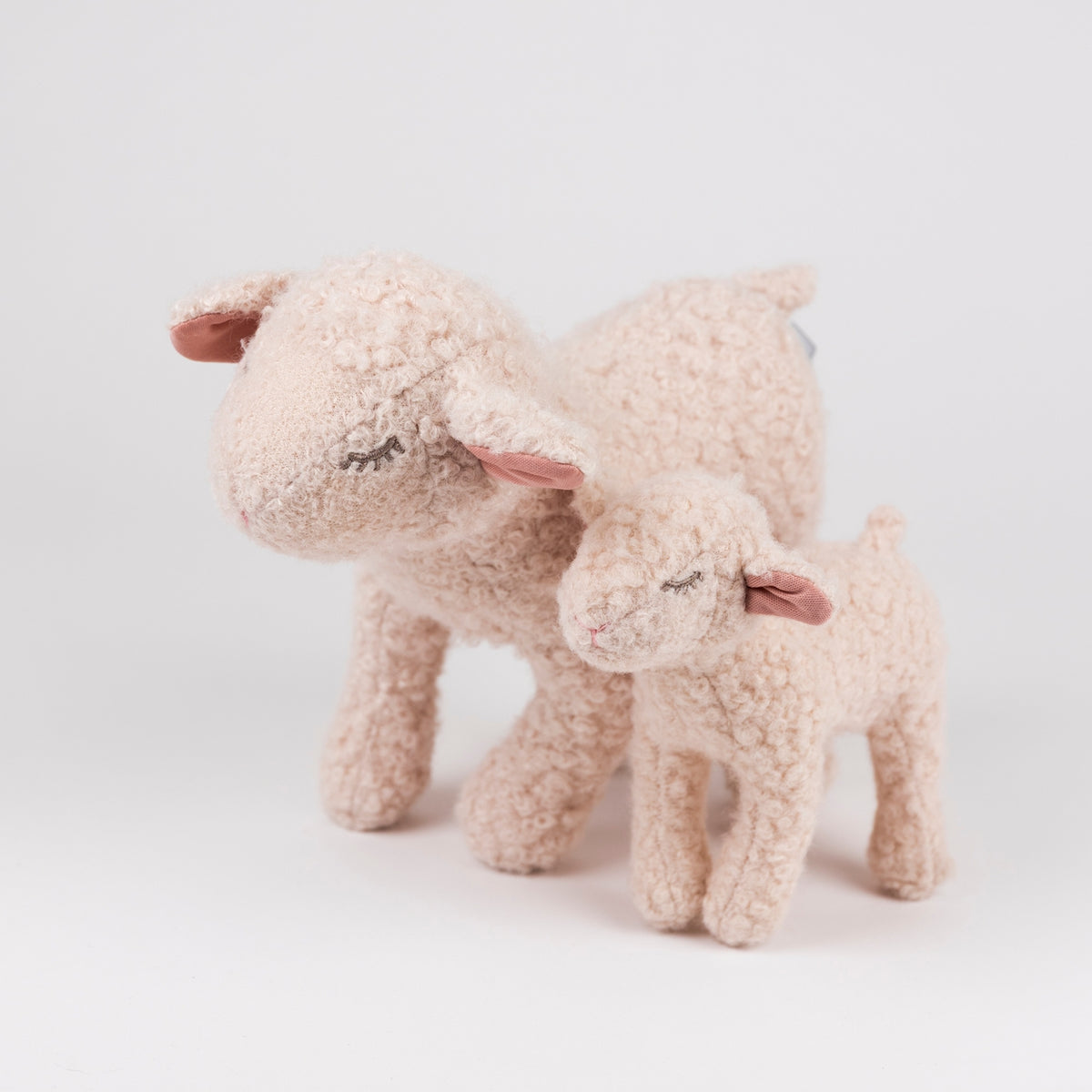 Two plush sheep toys, one larger adult sheep and one smaller lamb, in a soft cream-beige bouclé fabric with pink inner ears, photographed on a clean white studio background.