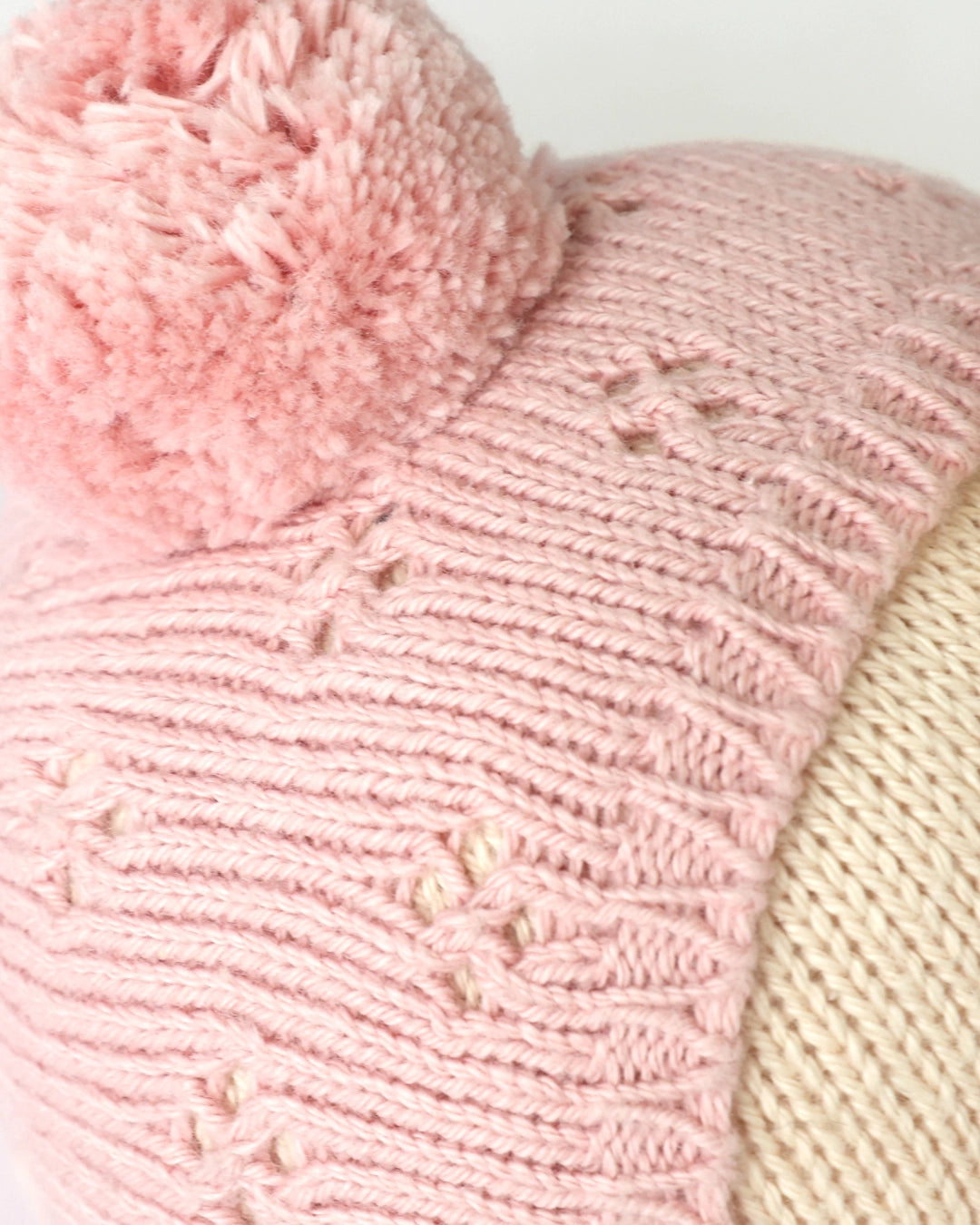 A close-up of a pink knitted beanie hat with a fluffy pom-pom on top and a ribbed cream-colored side panel.