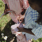 Two children outdoors on grass holding and playing with pastel-colored plastic toy gears and wheels. One child wears a pink-and-white gingham dress, and the other wears a blue-and-white gingham top.