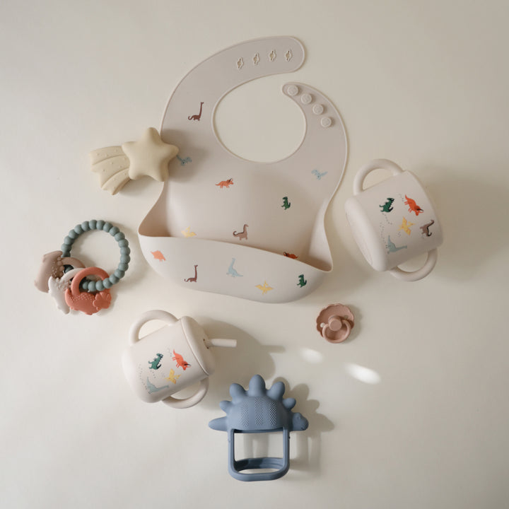 Collection of baby accessories including a bib, cups, and teething toys on a light background