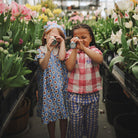 Two young children standing outdoors among blooming flowers and greenery, each holding up a Mushie kaleidoscope to one eye.