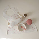 Baby bib, mat, and cup set with pink accents on a light background
